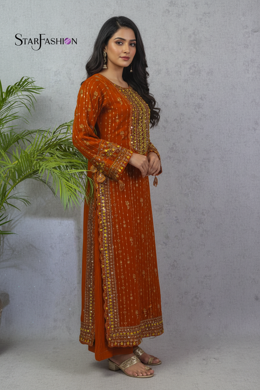 A woman modeling a rust-orange long kurta with gold and silver embroidery, standing against a textured gray wall with a green plant on the left. The brand logo 'STARFASHION' is visible in the top left corner.