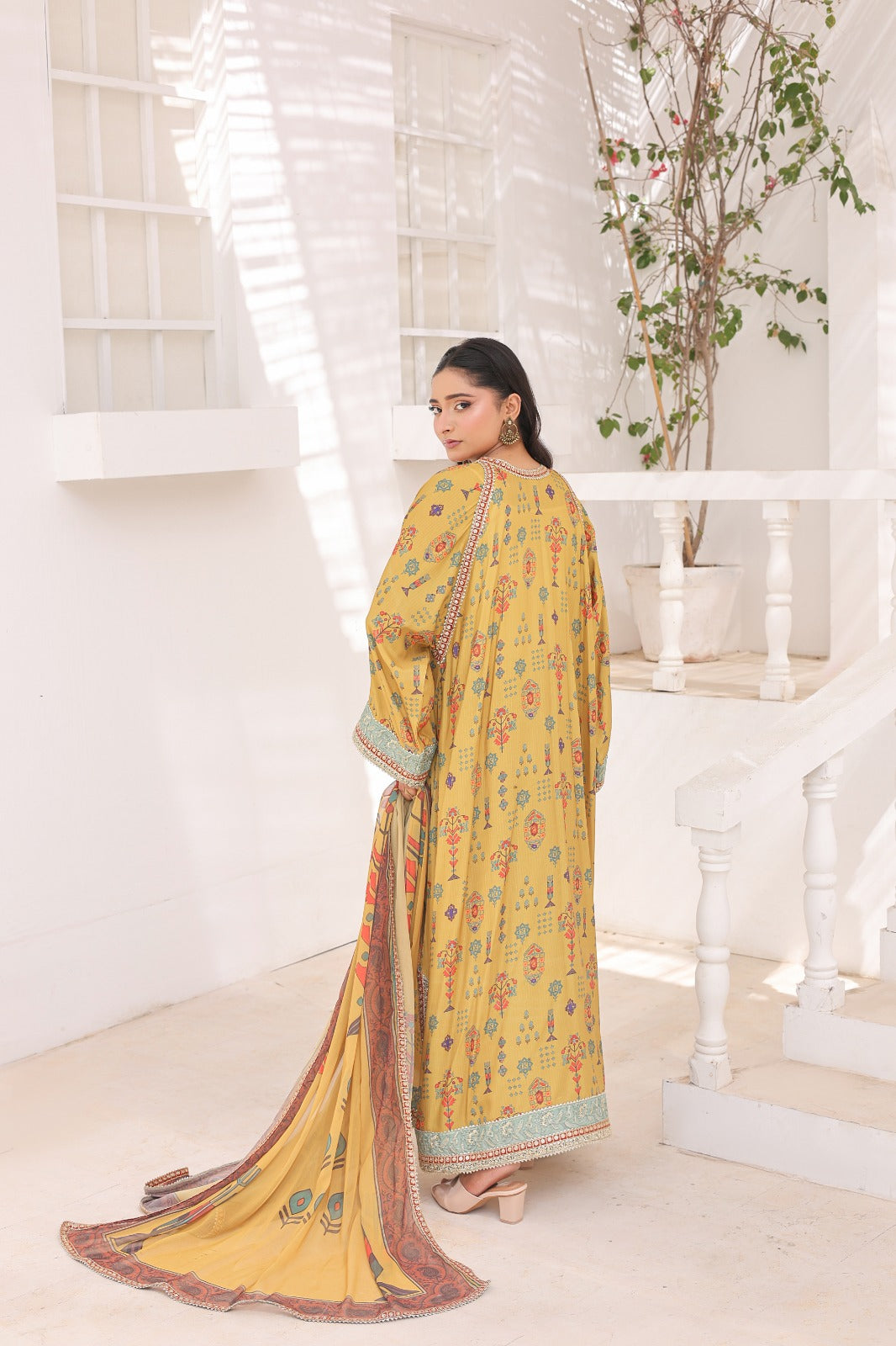 A woman wearing a yellow or mustard printed traditional outfit with a long dupatta, standing in a bright white interior with stairs and a potted plant in the background.