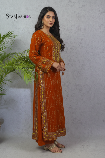 A woman modeling a rust-orange long kurta with gold and silver embroidery, standing against a textured gray wall with a green plant on the left. The brand logo 'STARFASHION' is visible in the top left corner.