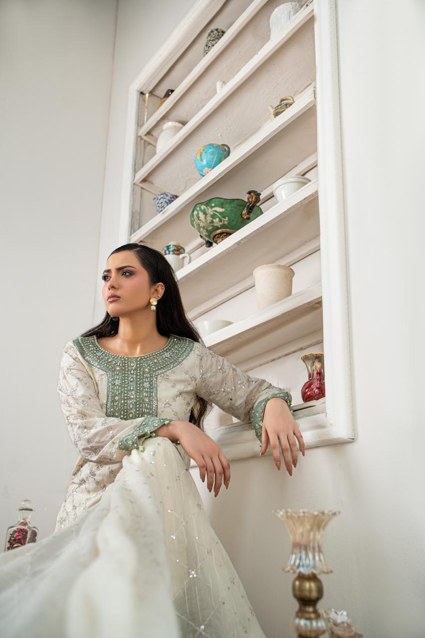 A woman wearing a traditional embroidered white and green Anarkali gown sits on a white shelf in a decorated room.