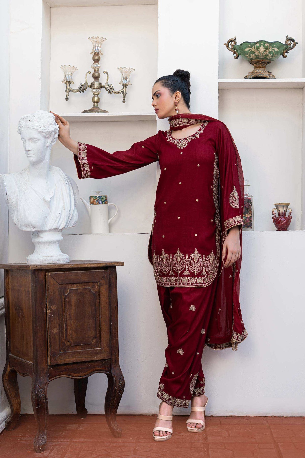 A woman models a deep maroon red traditional South Asian shalwar kameez suit with gold embroidery, standing in a styled room with a white bust sculpture and wooden table.