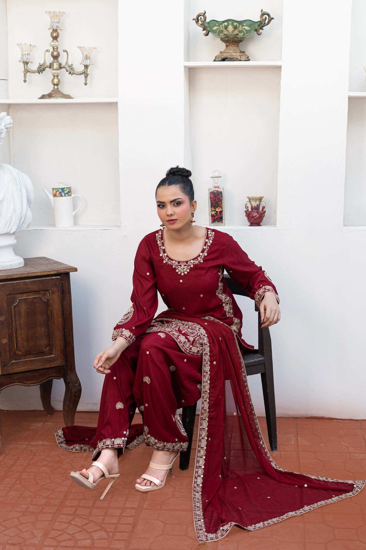 A woman sits on a black chair wearing a deep maroon traditional ethnic outfit with gold embroidery and a matching dupatta, set against a white wall with built-in shelves. Traditional Party Wear