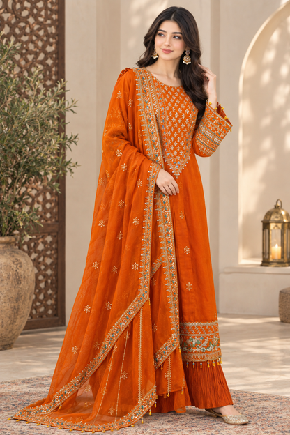 A woman models a traditional orange georgette salwar kameez suit with intricate gold and silver embroidery, featuring a matching dupatta, in an elegant indoor setting with warm architectural details.