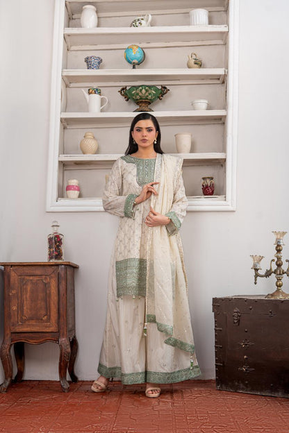 A woman stands in a room wearing a traditional cream-colored three-piece outfit with sage green borders and a matching dupatta. The setting features a white wall with built-in shelves holding decorative items, a wooden side table, and a dark brown trunk.