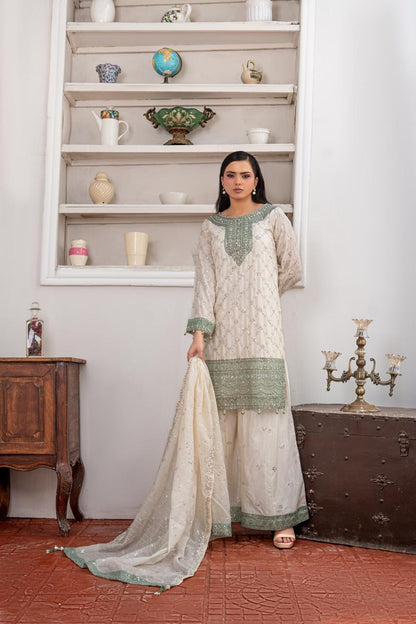 A woman models a traditional South Asian three-piece shalwar kameez suit in cream with sage green embroidery and borders, paired with a matching sheer dupatta, set against a vintage-style room background with wooden furniture and shelves.