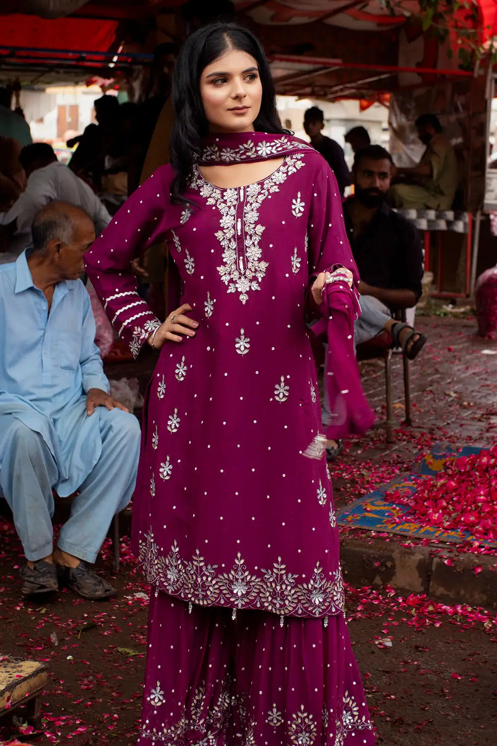 A woman modeling a deep magenta purple ethnic salwar kameez suit with intricate silver mirror work embroidery, standing in an outdoor market setting with people and flower petals on the ground.