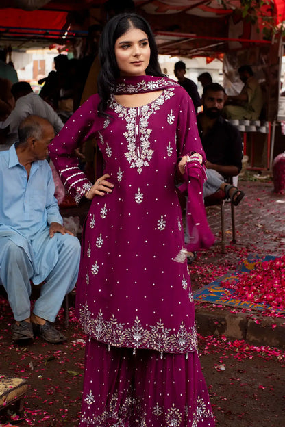A woman modeling a deep magenta purple ethnic salwar kameez suit with intricate silver mirror work embroidery, standing in an outdoor market setting with people and flower petals on the ground.