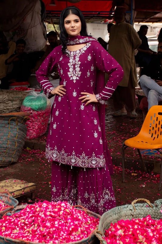 A woman stands in a market setting wearing a deep magenta purple traditional outfit with intricate silver embroidery and white floral patterns. The outfit consists of a long kurta, matching pants, and a dupatta.