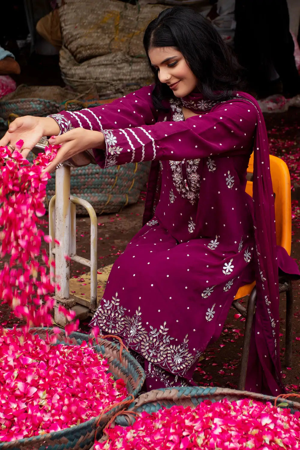 A woman wearing a deep magenta purple traditional salwar kameez suit with white floral embroidery and silver mirror work, sitting in an orange chair amidst baskets of bright pink rose petals.