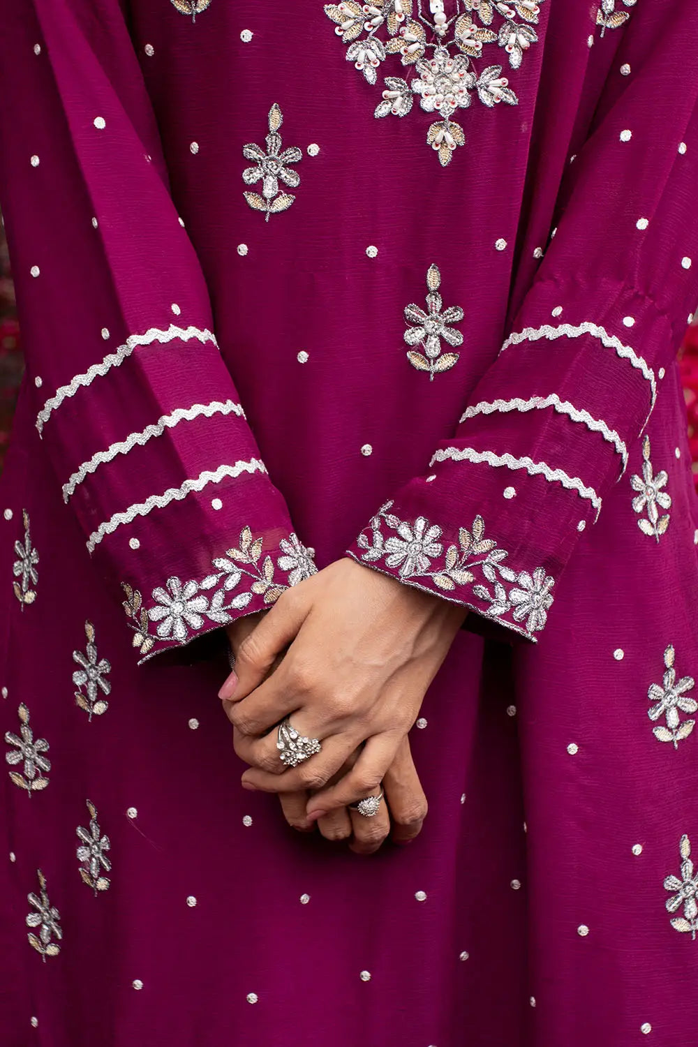 A close-up of a embroided magenta purple traditional dress with intricate silver floral embroidery and white polka dots, featuring a person's hands clasped in the center.
