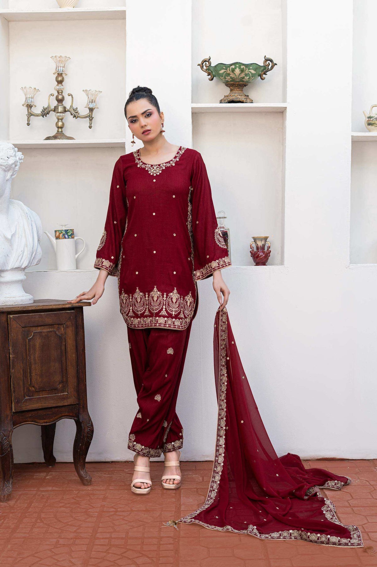 A woman models a deep maroon three-piece salwar kameez suit with gold embroidery, featuring a short kurta, matching trousers, and a draped dupatta, posed in front of a white wall with built-in shelves.