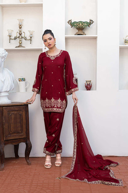A woman models a deep maroon three-piece salwar kameez suit with gold embroidery, featuring a short kurta, matching trousers, and a draped dupatta, posed in front of a white wall with built-in shelves.