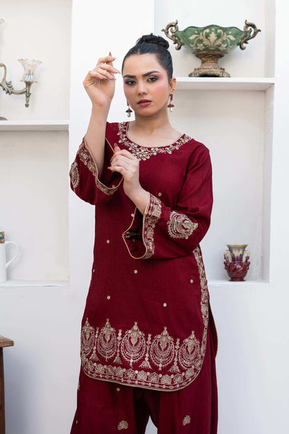 A woman modeling a deep maroon traditional three-piece salwar kameez suit with intricate gold embroidery, standing in front of a white wall with built-in shelves.