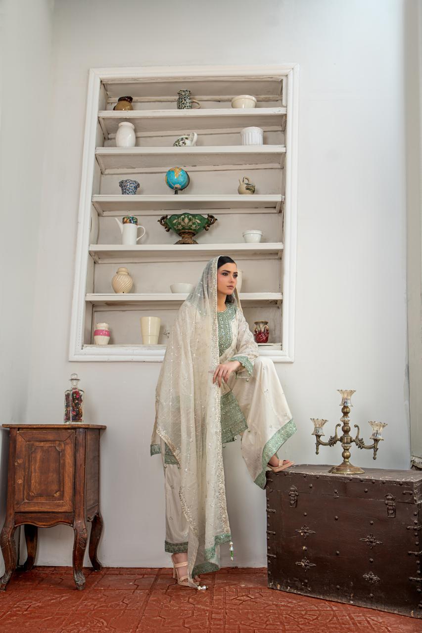 A woman models a traditional South Asian outfit consisting of a light beige/cream kurta and dupatta with sage green borders, standing in a styled room with a built-in white shelf, a wooden cabinet, and a vintage trunk.