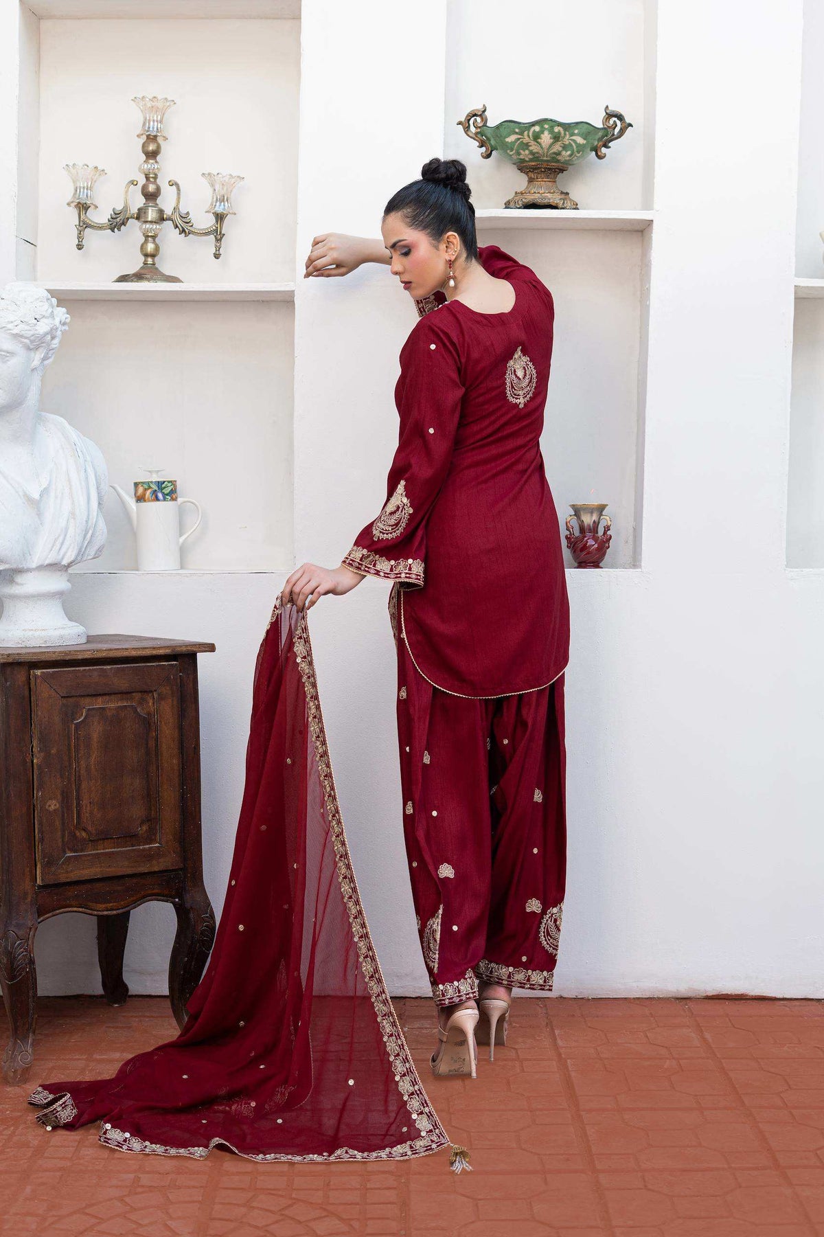 A woman models a deep maroon three-piece shalwar kameez suit with gold embroidery and a matching dupatta in an indoor setting with white walls and terracotta flooring.