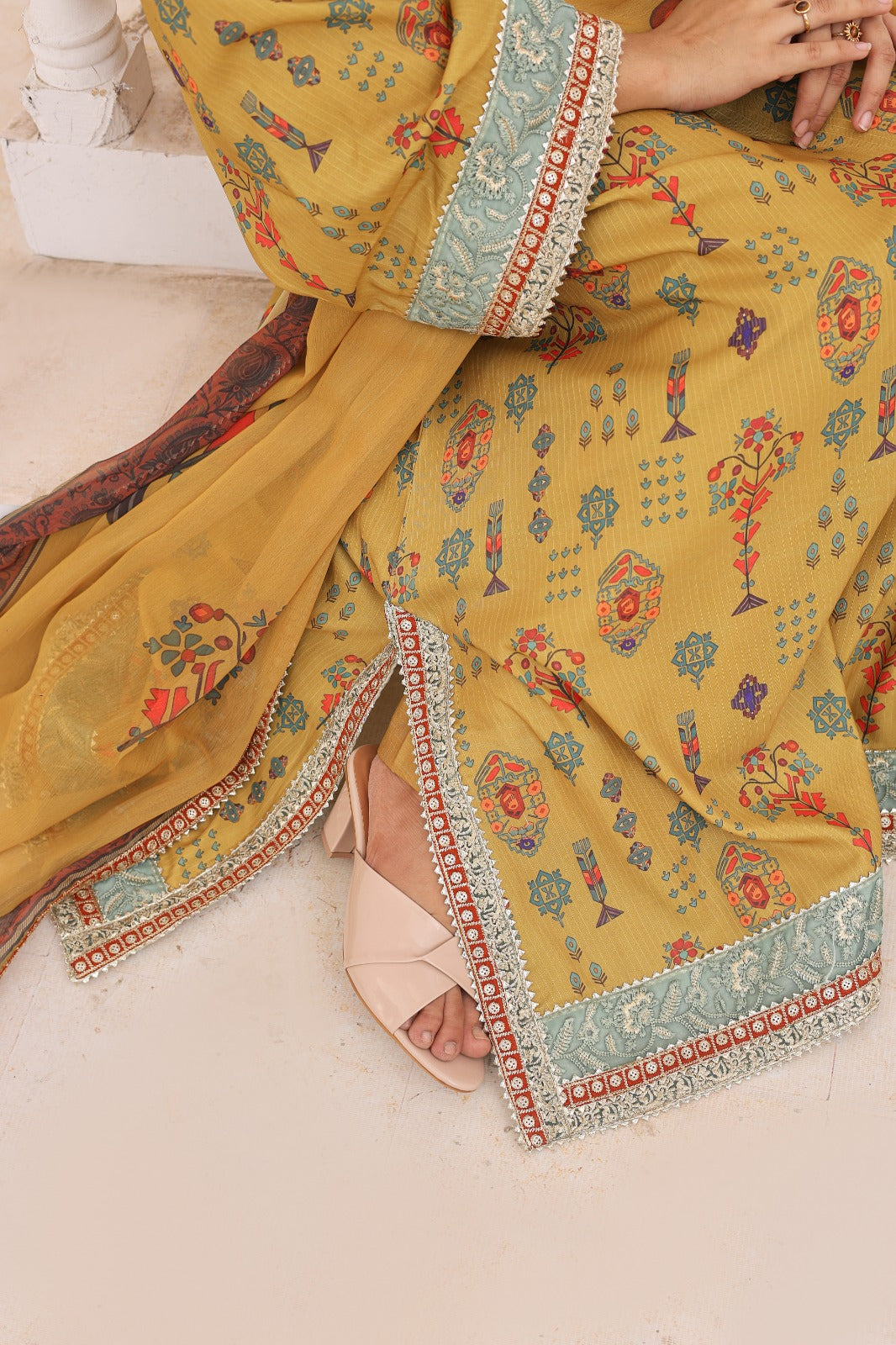 A close-up of a mustard yellow traditional outfit with colorful floral prints and ornate light blue borders, worn by a model seated on a light stone floor.