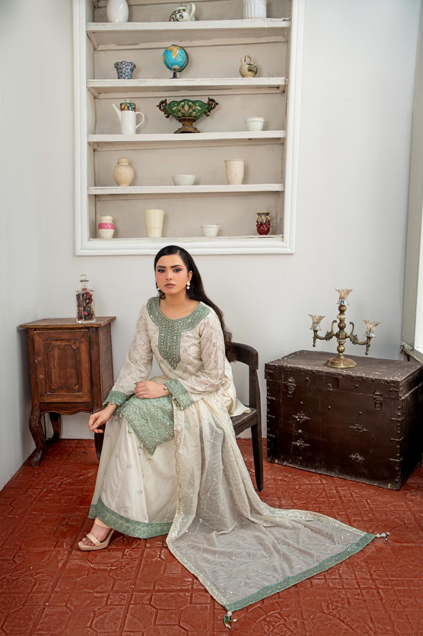 A woman sits on a dark wooden chair in a styled room, wearing a traditional South Asian shalwar kameez outfit. The outfit consists of an off-white/cream kurta with green embroidery and a matching green border, paired with a light green dupatta.